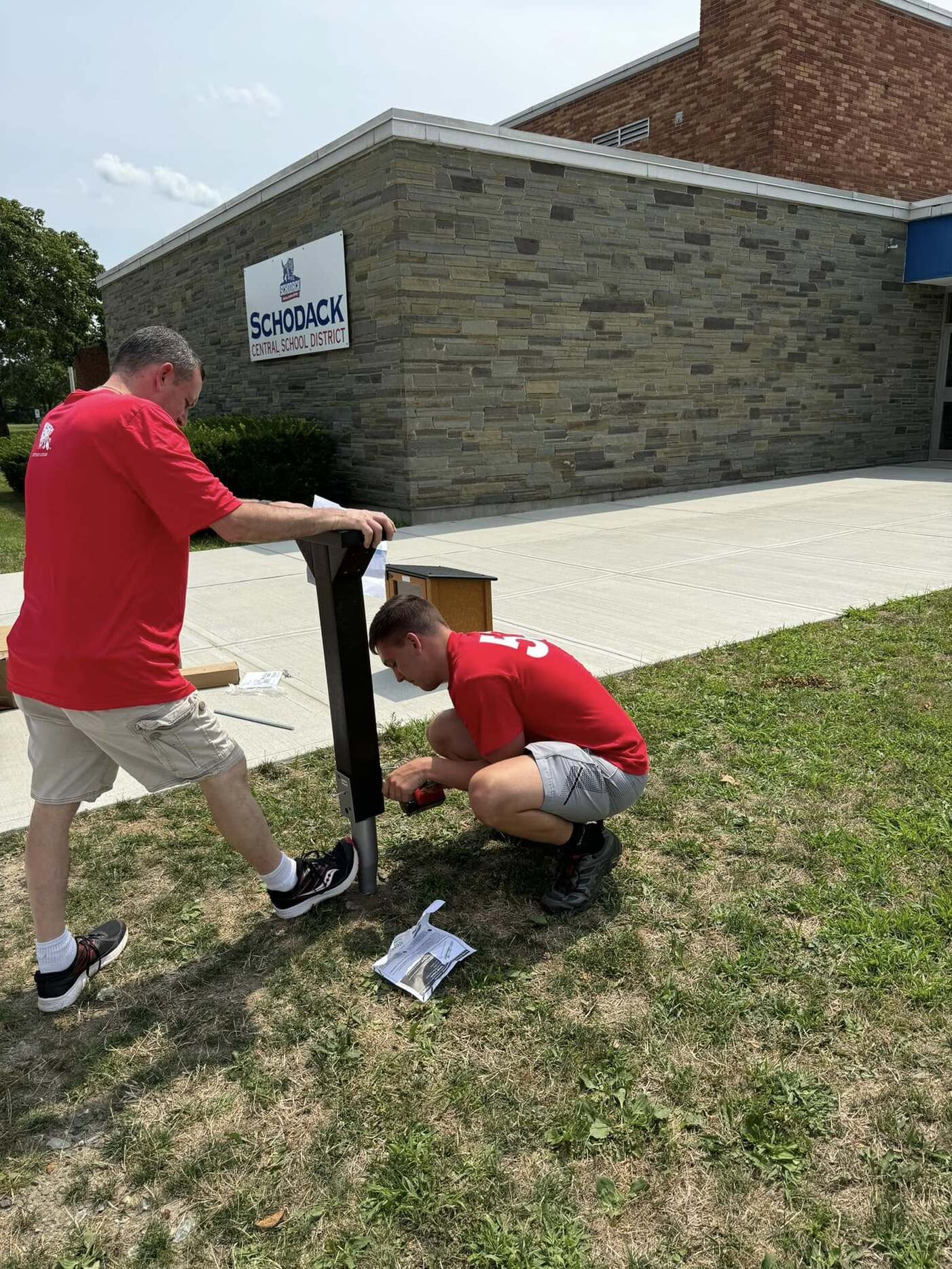 Two people in red shirts screwing a bolt into the bottom of a post.  One person is drilling, the other is holding the post steady.