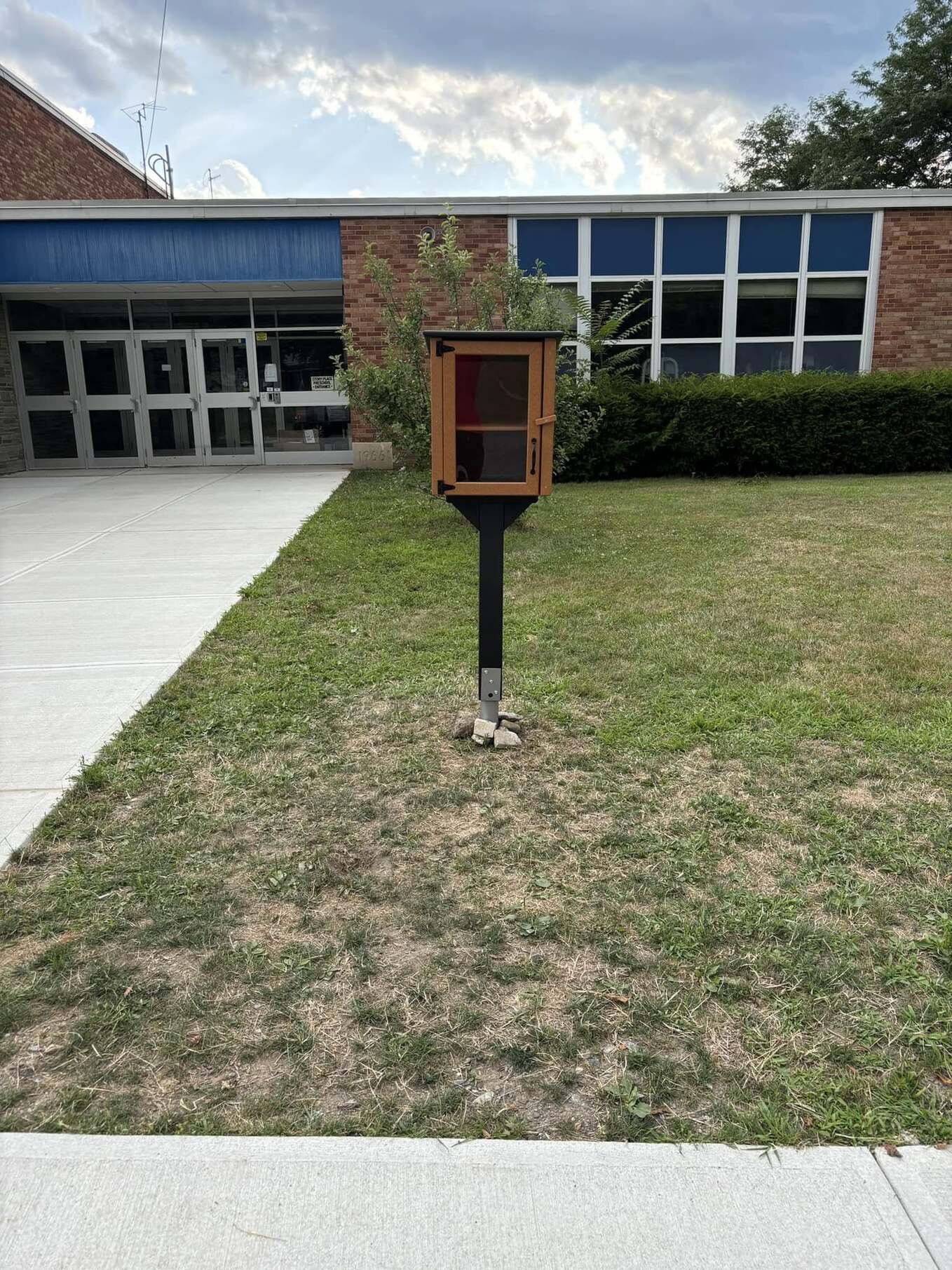 The little library to the right of a sidewalk outside of a brick building.