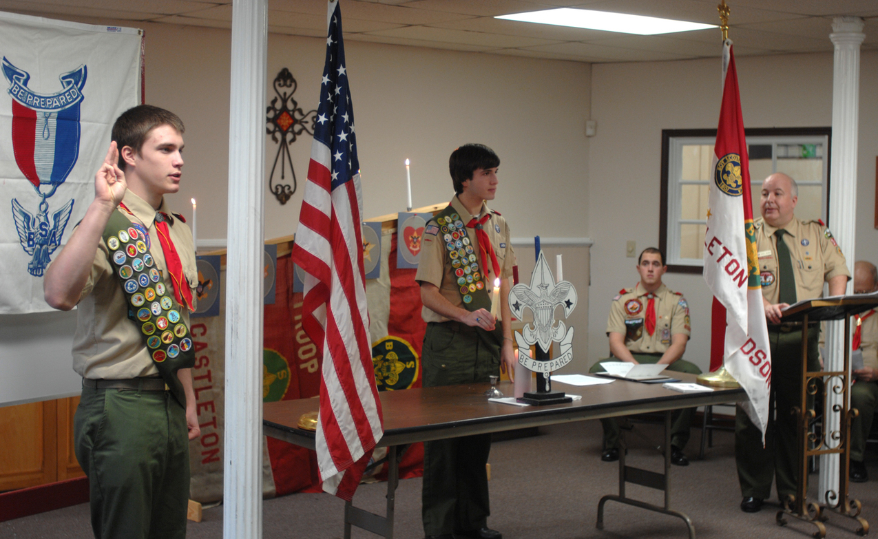 Four people wearing Class A Uniforms.  On the left is a scout giving the scout sign.  In the middle is a scout lighting a candle.  On the right is an adult speaking behind a podium.