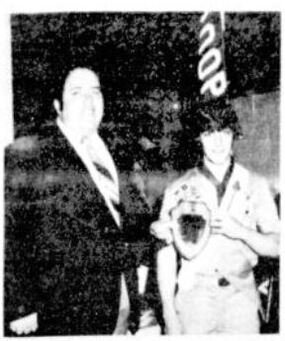 Two people.  The person on the left is wearing a suit, and presenting an award.  The person on the right is wearing a class A, an Order of the Arrow sash, and a merit badge sash.