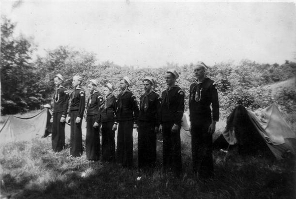 Eight Sea Scouts stand at attention in front of tents.  They are in their uniforms.  The photo is black-and-white.