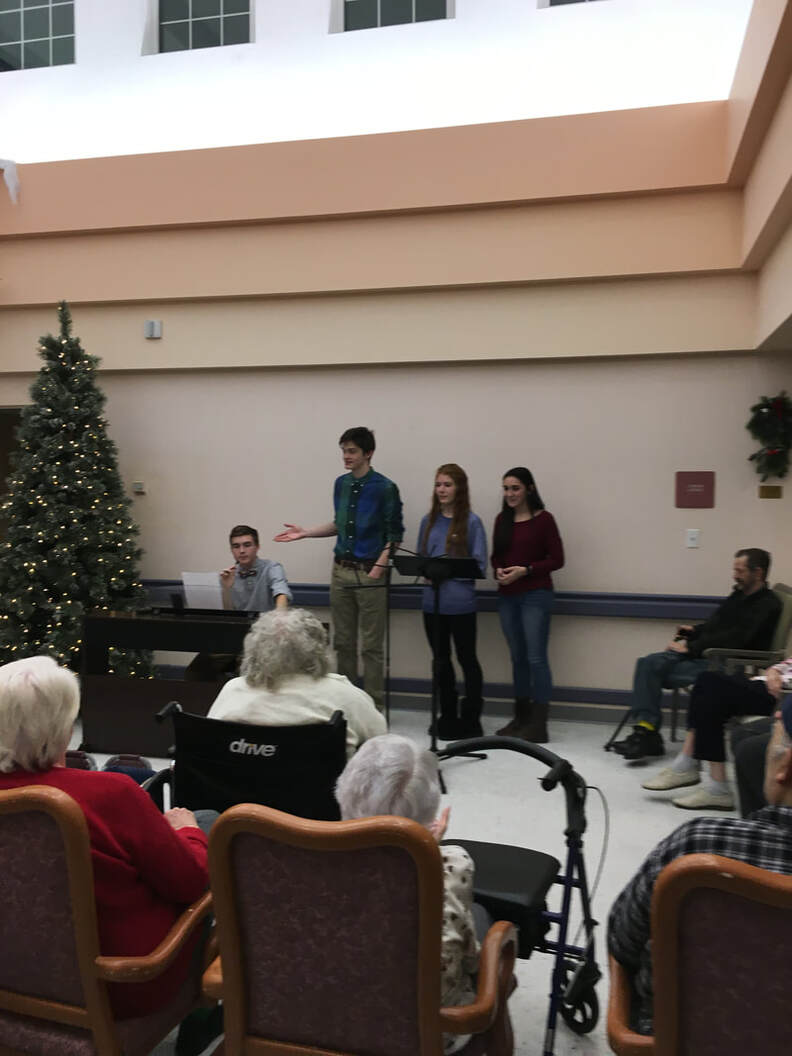 Seth is sitting behind a piano talking into a microphone.  There is a boy and 2 girl students to the right of him standing.