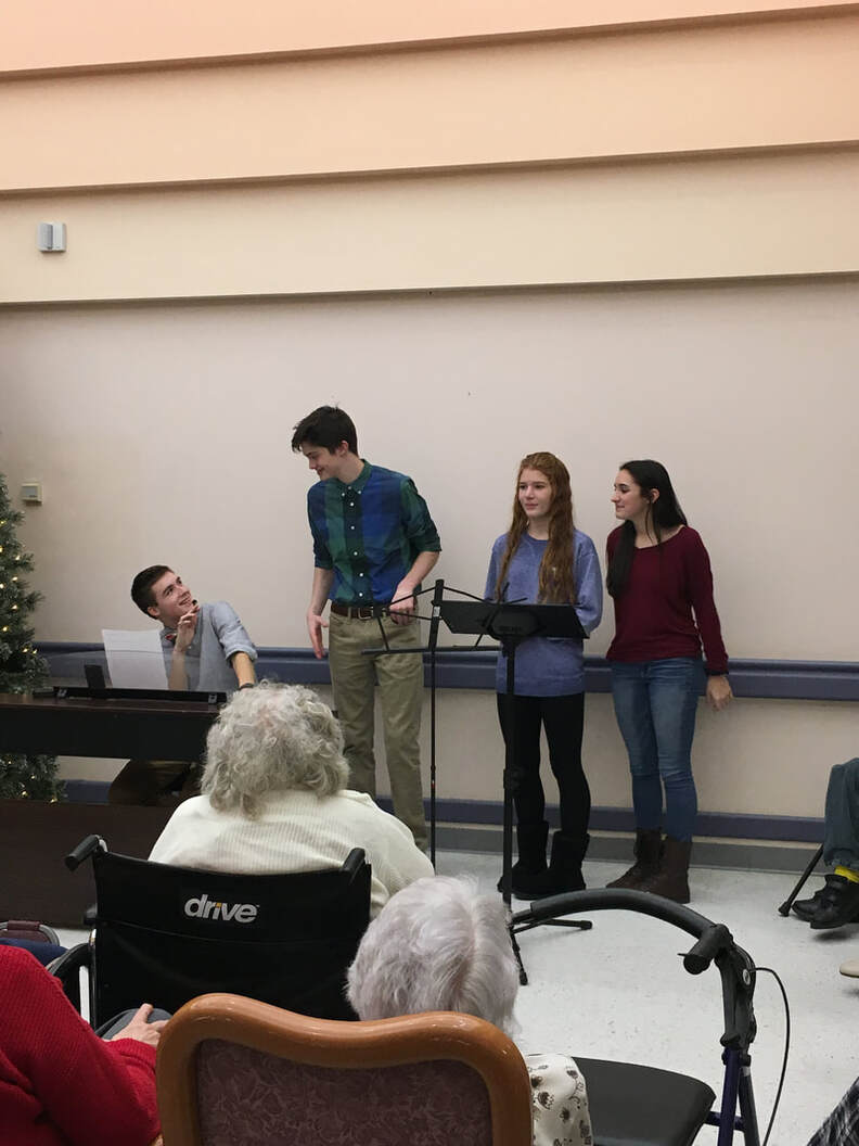 Seth is sitting behind a piano talking into a microphone.  There is a boy and 2 girl students to the right of him standing.