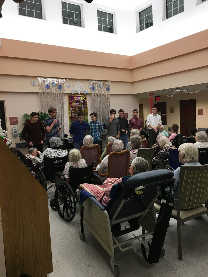 A group of students stand in front of senior citizens.  One of whom is talking to a microphone.