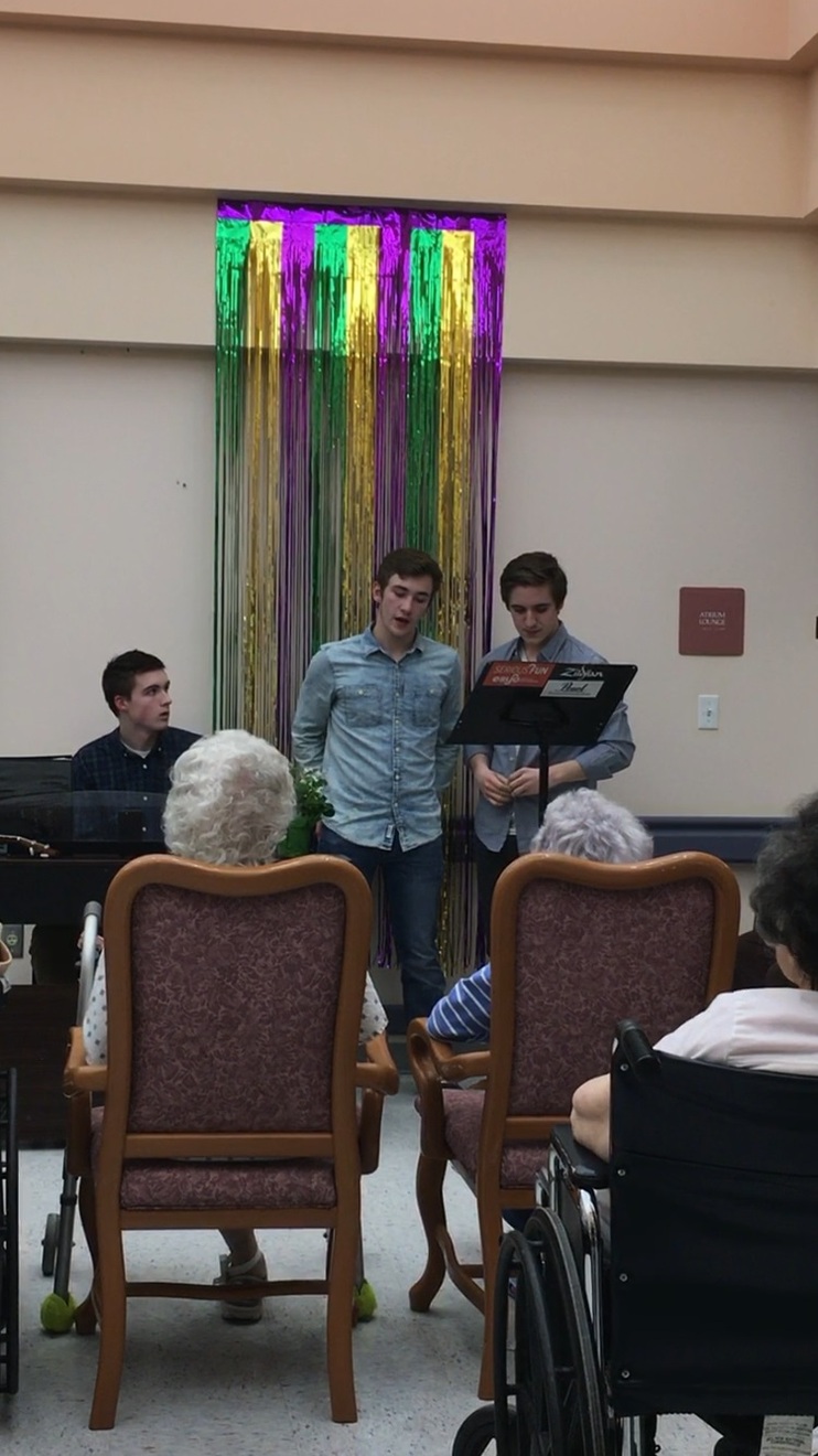 A student is sitting at a piano, while two male students stand to the right of him, singing.