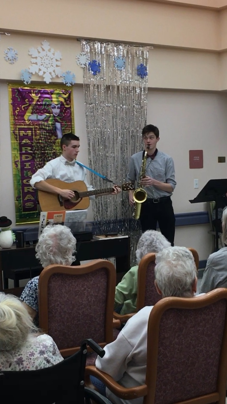 The student on the left playing an acoustic guitar, the student on the right playing a saxophone.