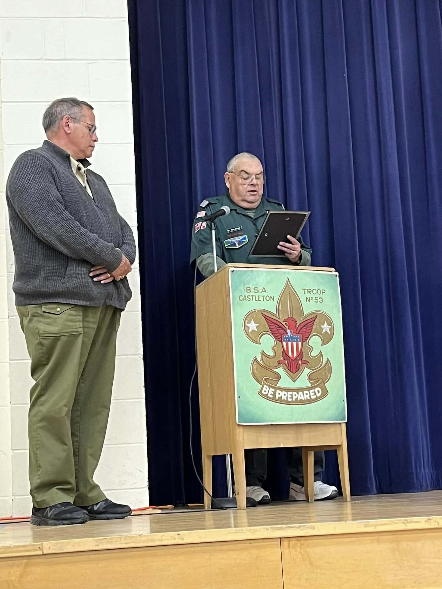 Two men stand behind a podium.  One is wearing a green class A and speaking.