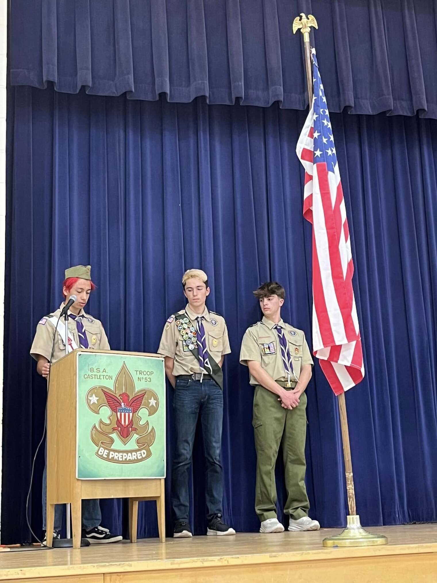 A three scouts in tan class A uniforms speaking into a microphone at a podium.