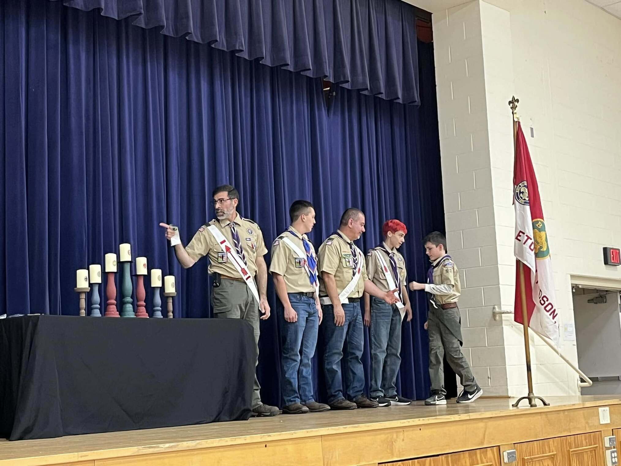 Five scouts and adults standing on stage.  The scoutmaster is pointing to the left (his right).