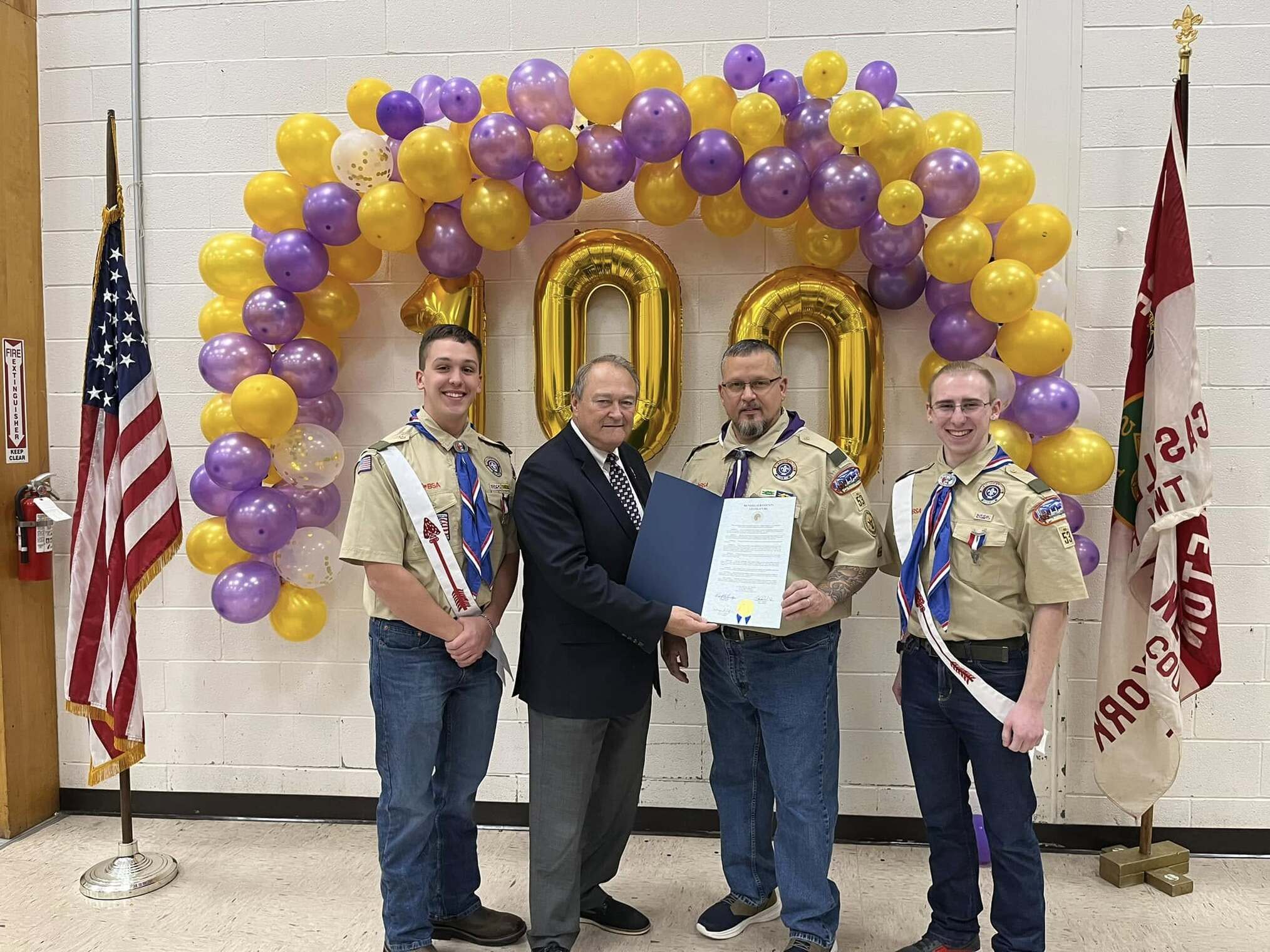 Three people in class A uniforms and one in a suit stand in front of the balloon wall.  They are holding a proclamation.