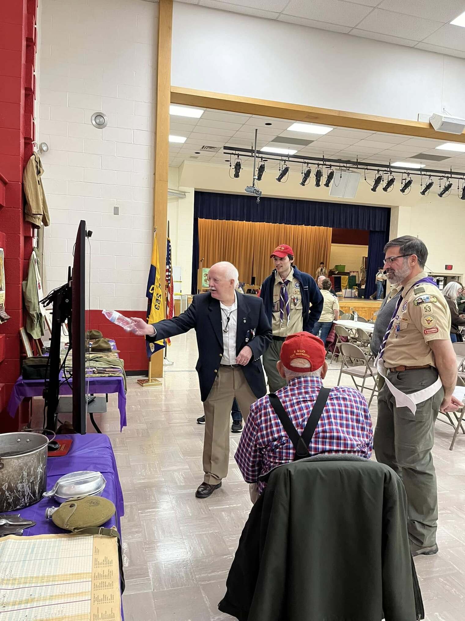 A man in a suit jacket and an Eagle Scout metal points to the TV with a water bottle.